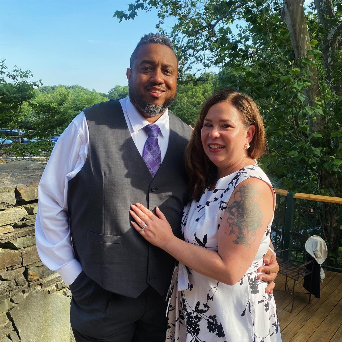 A smiling man in a grey vest and a woman in a floral white dress posing outdoors.