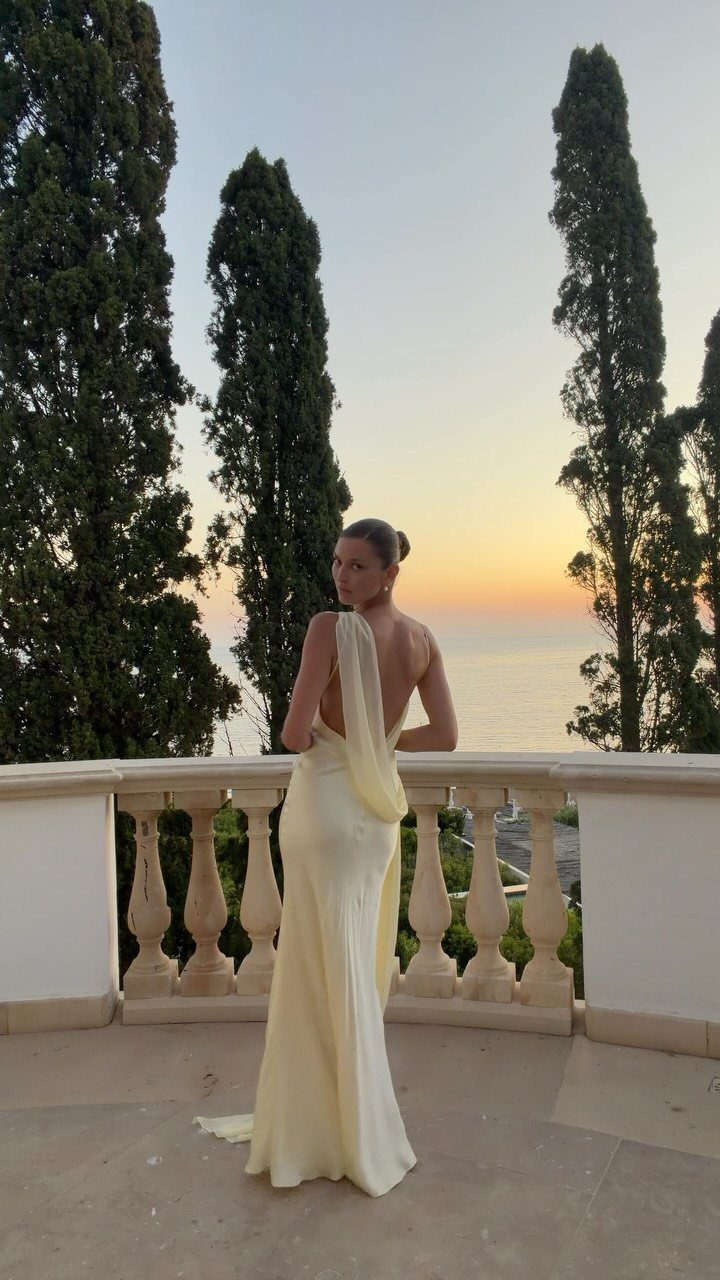 Woman in a cream backless formal gown standing on a balcony overlooking the ocean at sunset.
