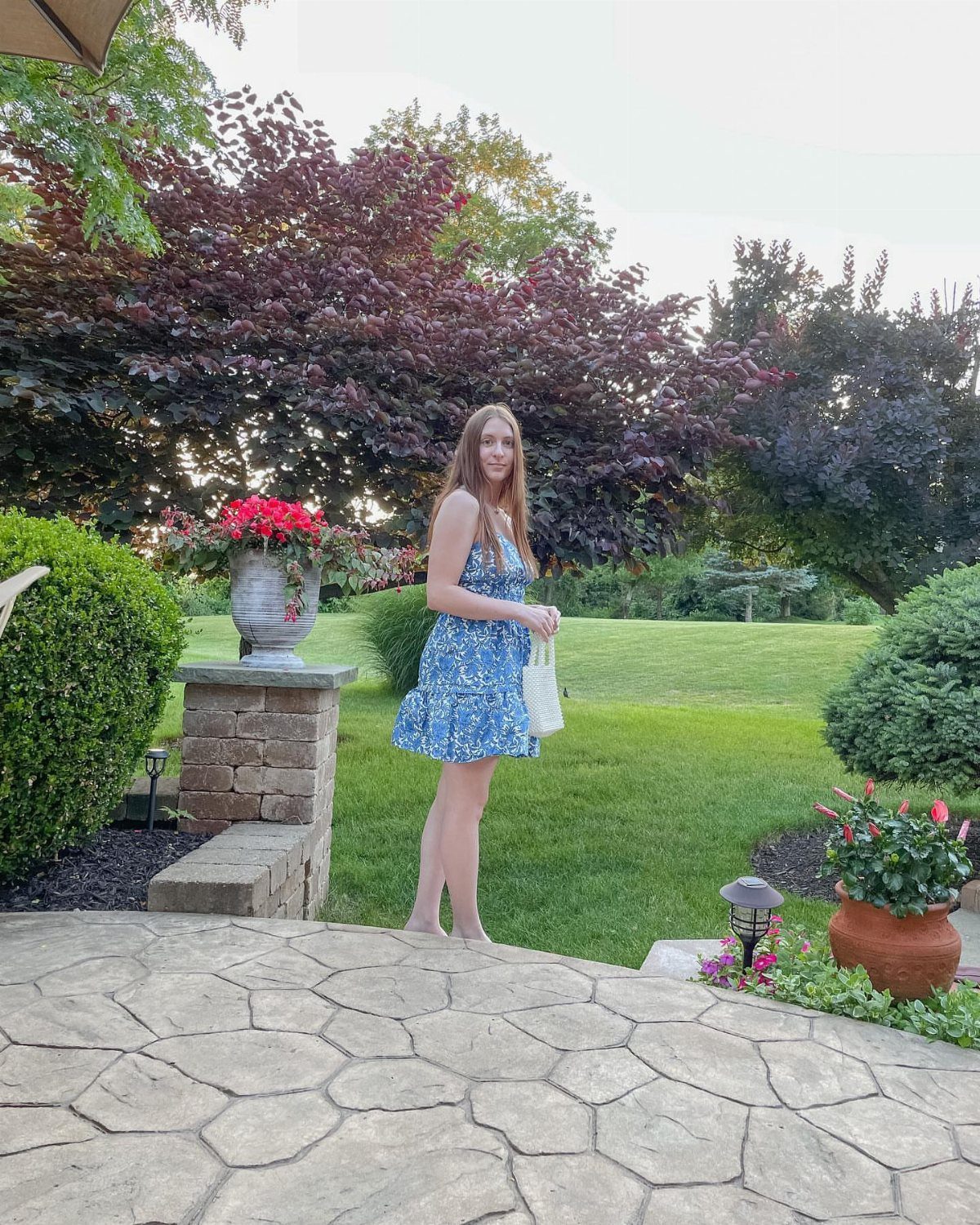 A young woman in a blue floral sundress stands on a stone patio in a lush garden.