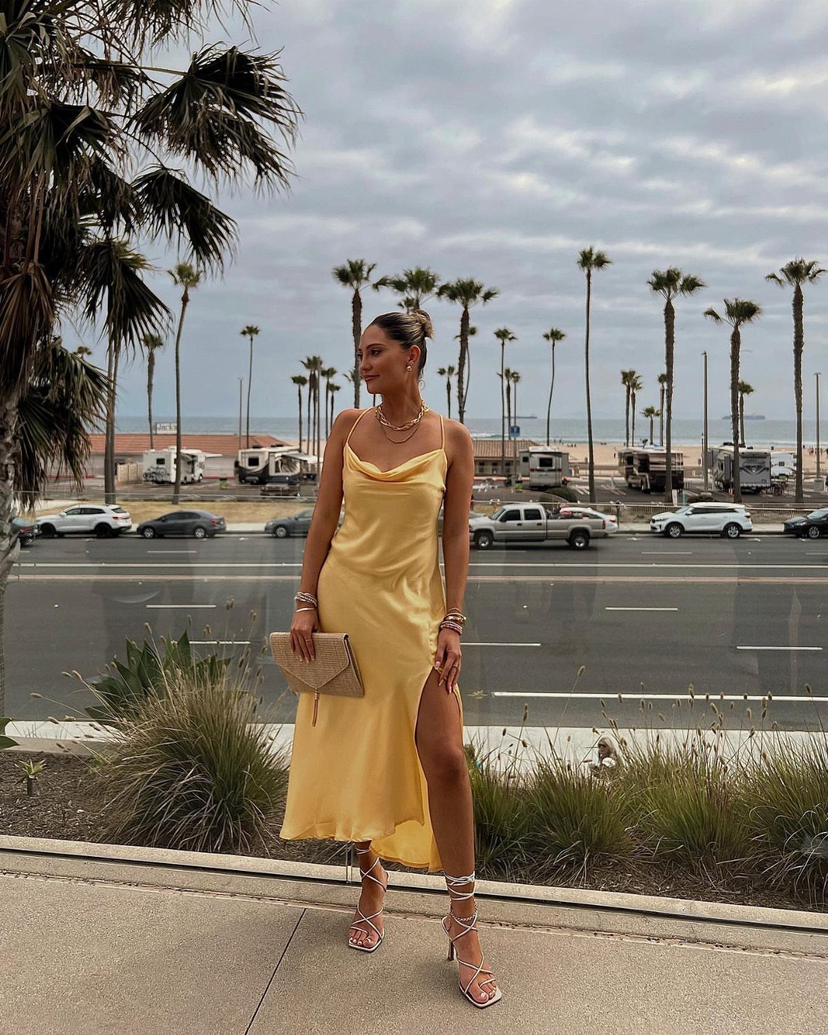 Woman in a long yellow slip dress and strappy sandals standing near palm trees.