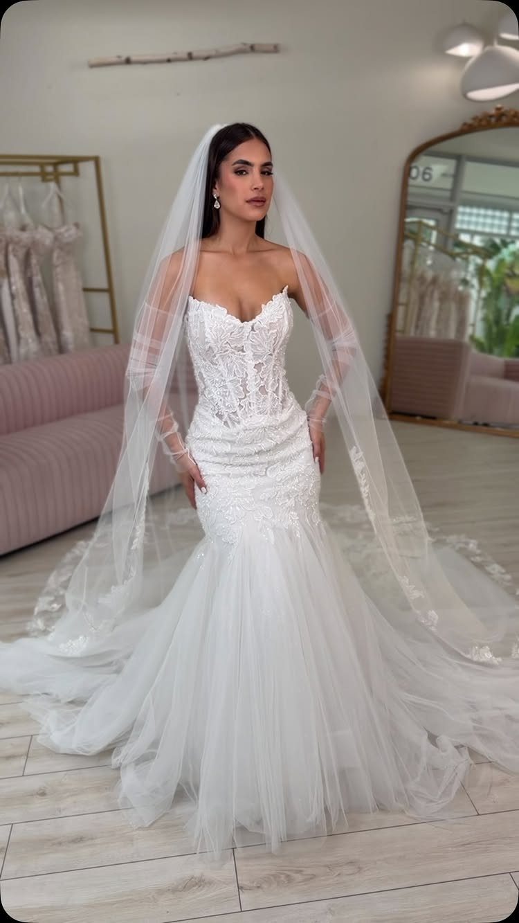 Woman in a white lace mermaid wedding dress and long veil posing in a boutique.
