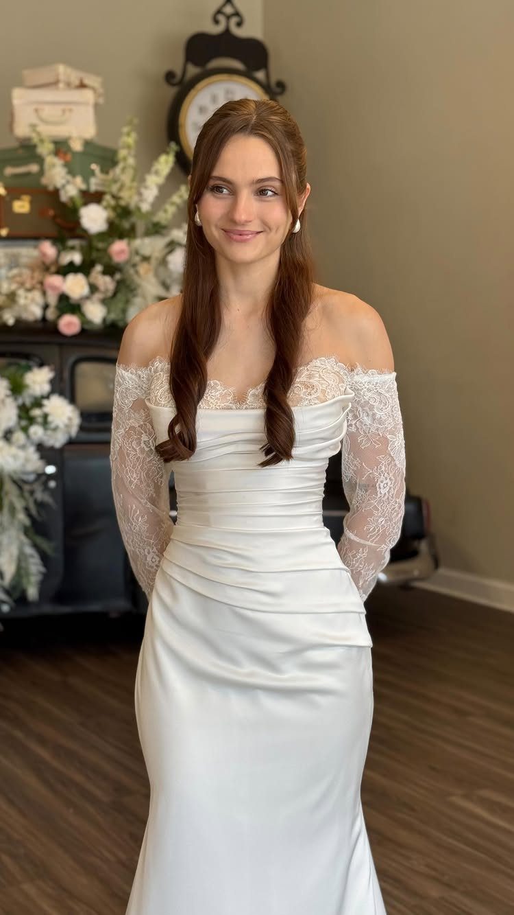 A woman smiling in an off-the-shoulder white wedding dress with long lace sleeves.
