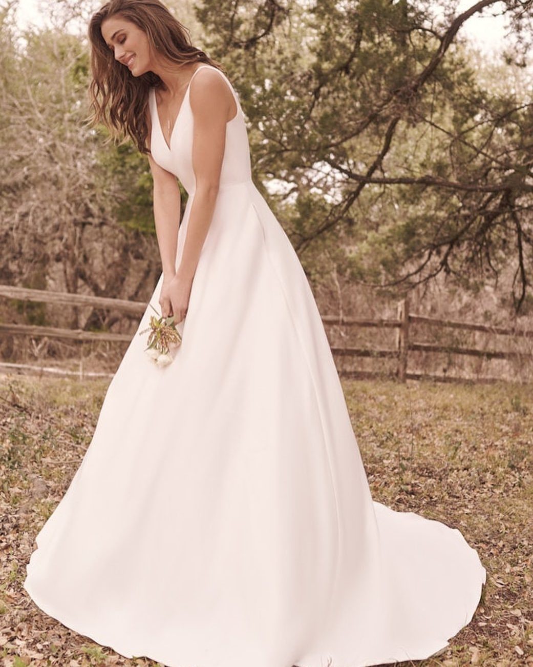 Woman wearing a minimalist white satin wedding gown standing in a grassy field with trees.