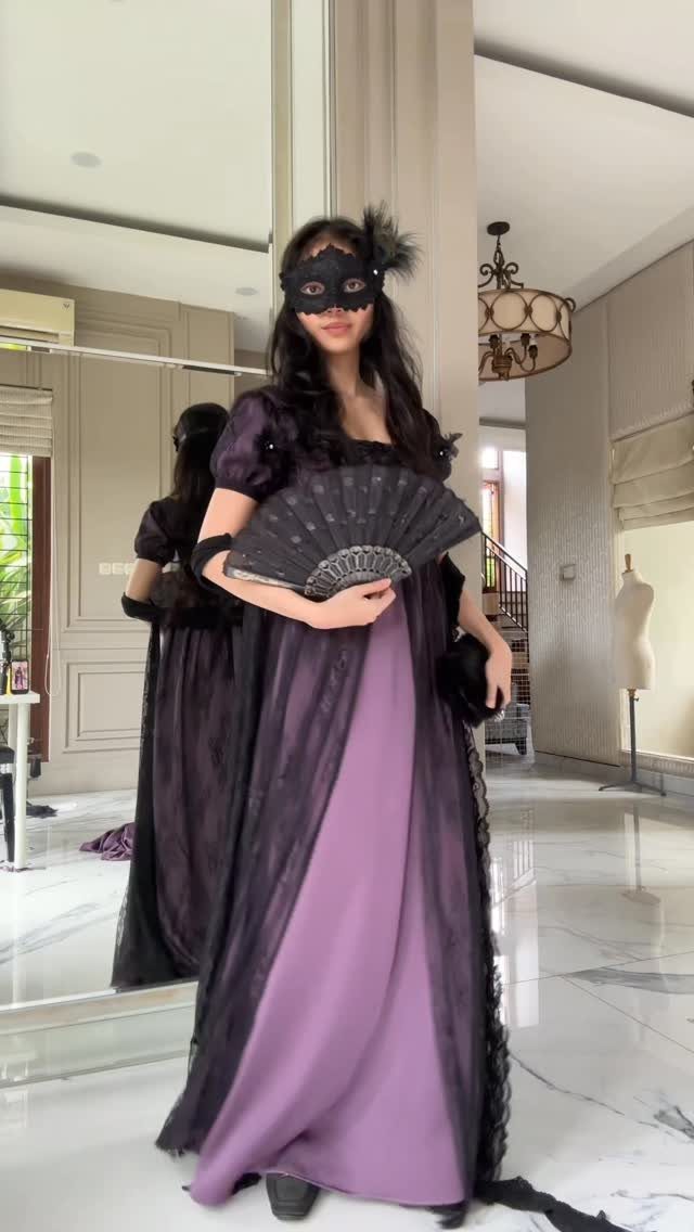Woman in a purple and black masquerade gown holding a black folding fan.