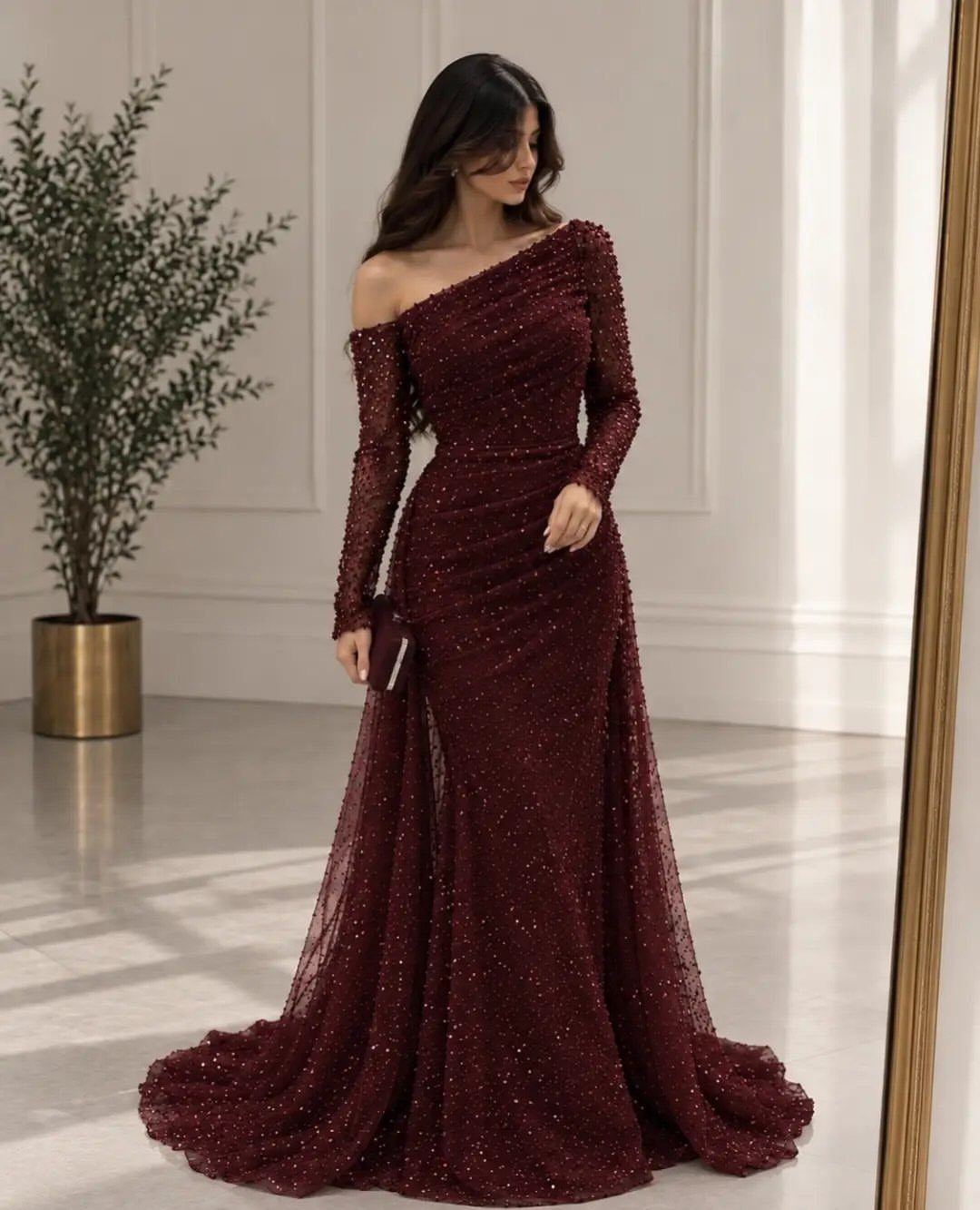 A woman wearing a sparkling burgundy one-shoulder evening gown standing in a bright room.