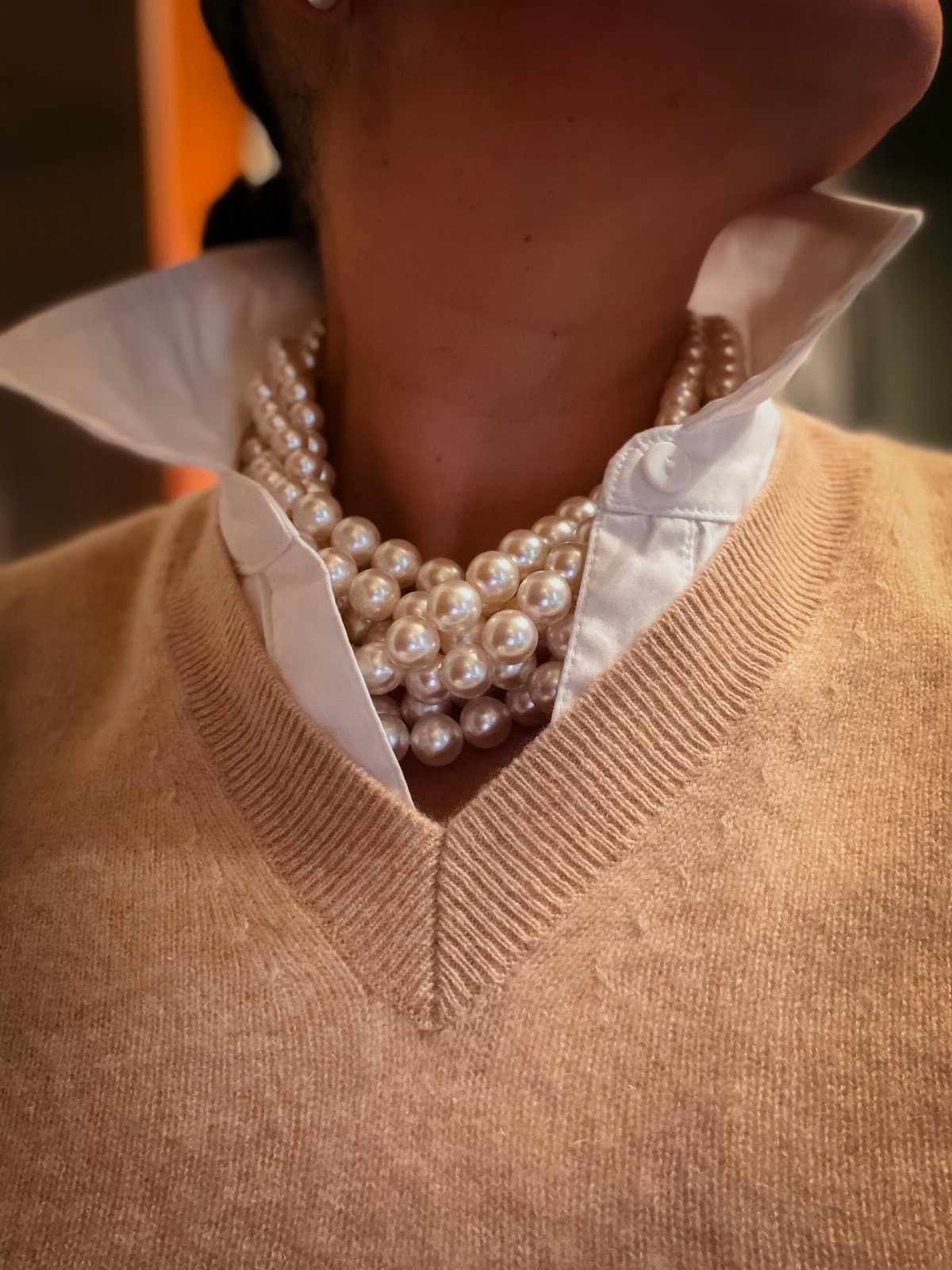 Person wearing a tan sweater, white collared shirt, and a multi-strand pearl necklace.