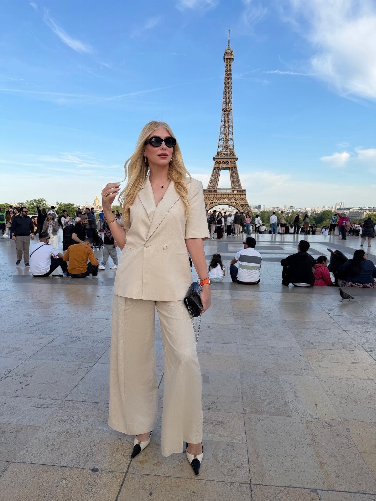Blonde woman in a beige coordinated suit standing in a plaza with the Eiffel Tower behind her.