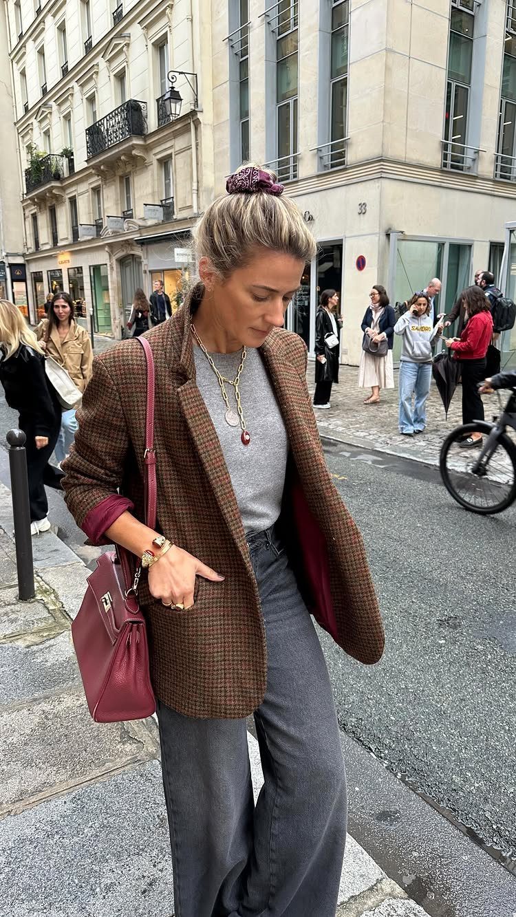 Woman in a brown blazer and grey top carrying a burgundy handbag on a city street.