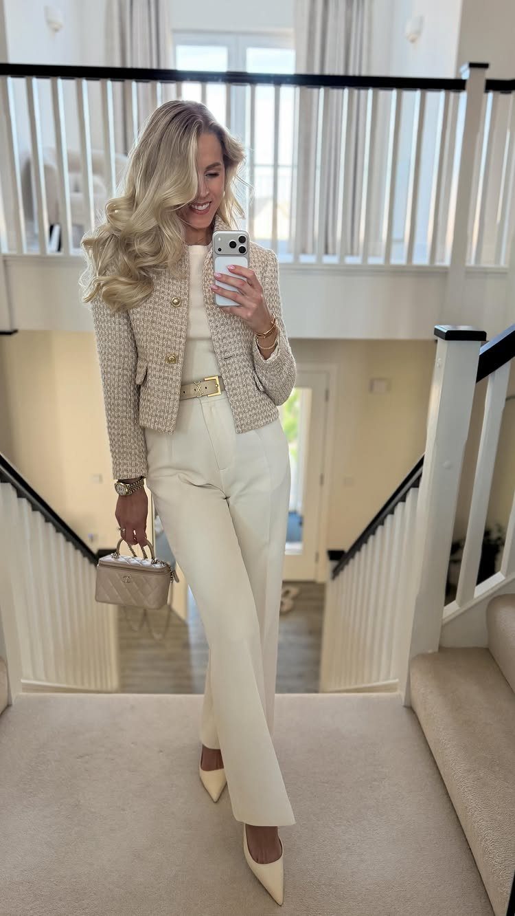 Blonde woman in cream tweed jacket and trousers posing for a mirror selfie on stairs.