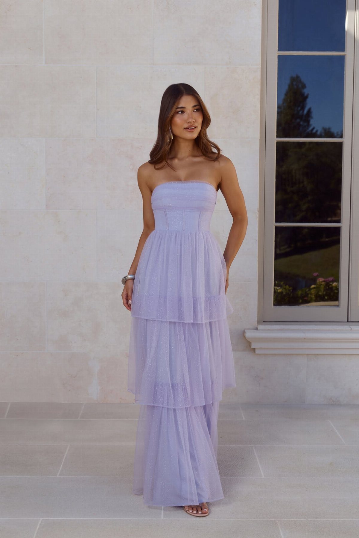 A woman posing in a strapless, tiered light purple tulle maxi dress against a stone wall.