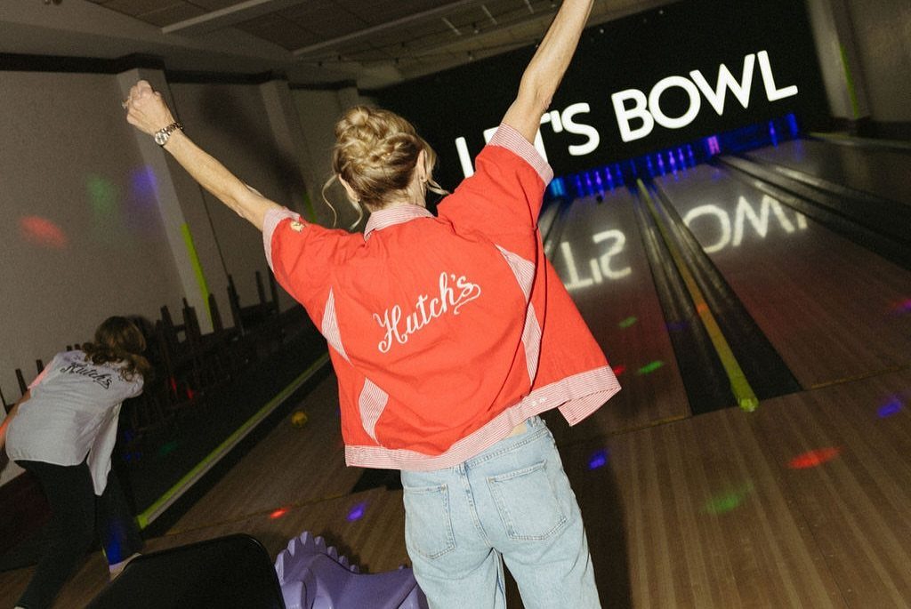 A woman in a red jacket celebrates with arms raised in a bowling alley.