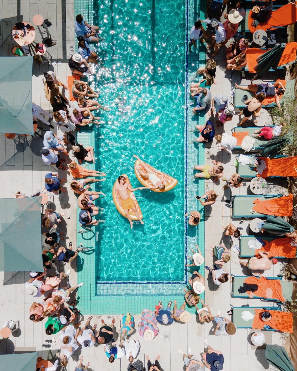 People lounging around a bright turquoise swimming pool with two individuals floating on yellow rafts.