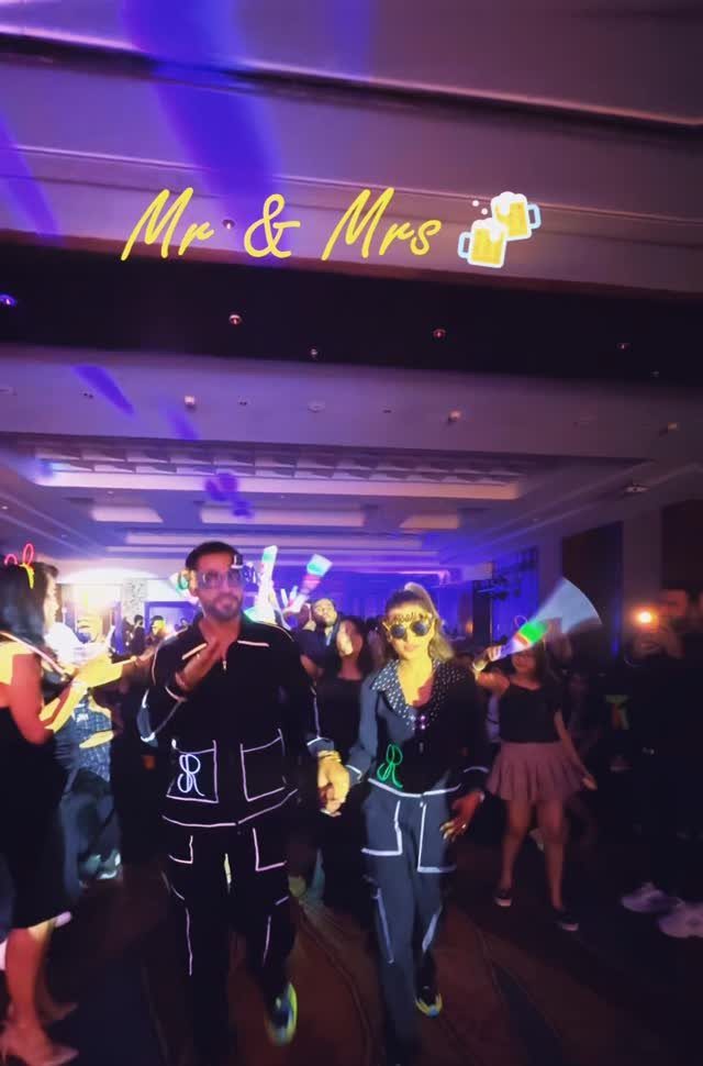 A couple wearing neon-lined black outfits walks through a dark, crowded dance floor with blue lighting.