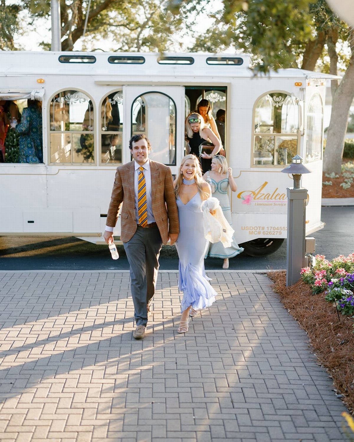 A dressed-up couple walks away from a white vintage shuttle bus parked on a brick path.
