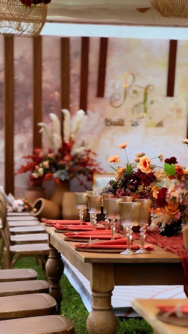 Wooden dining table set with red napkins, gold glassware, and vibrant floral arrangements outdoors.