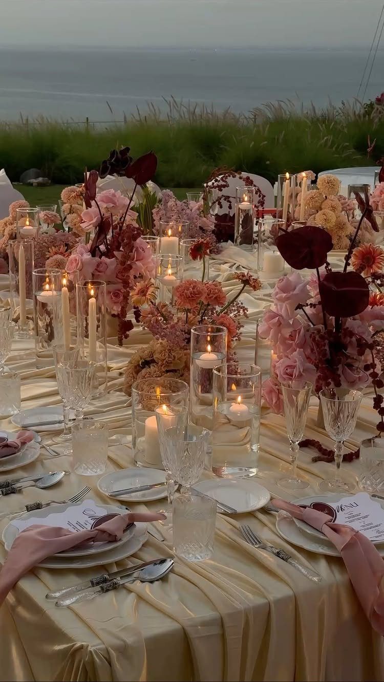 A formal wedding table setting with gold linens, pink flowers, and glowing candles by the sea.