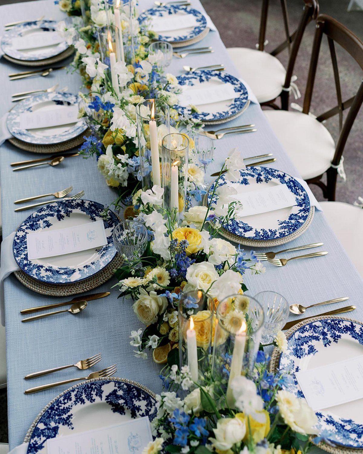 A long wedding table set with blue patterned plates, gold cutlery, and yellow and blue floral arrangements.