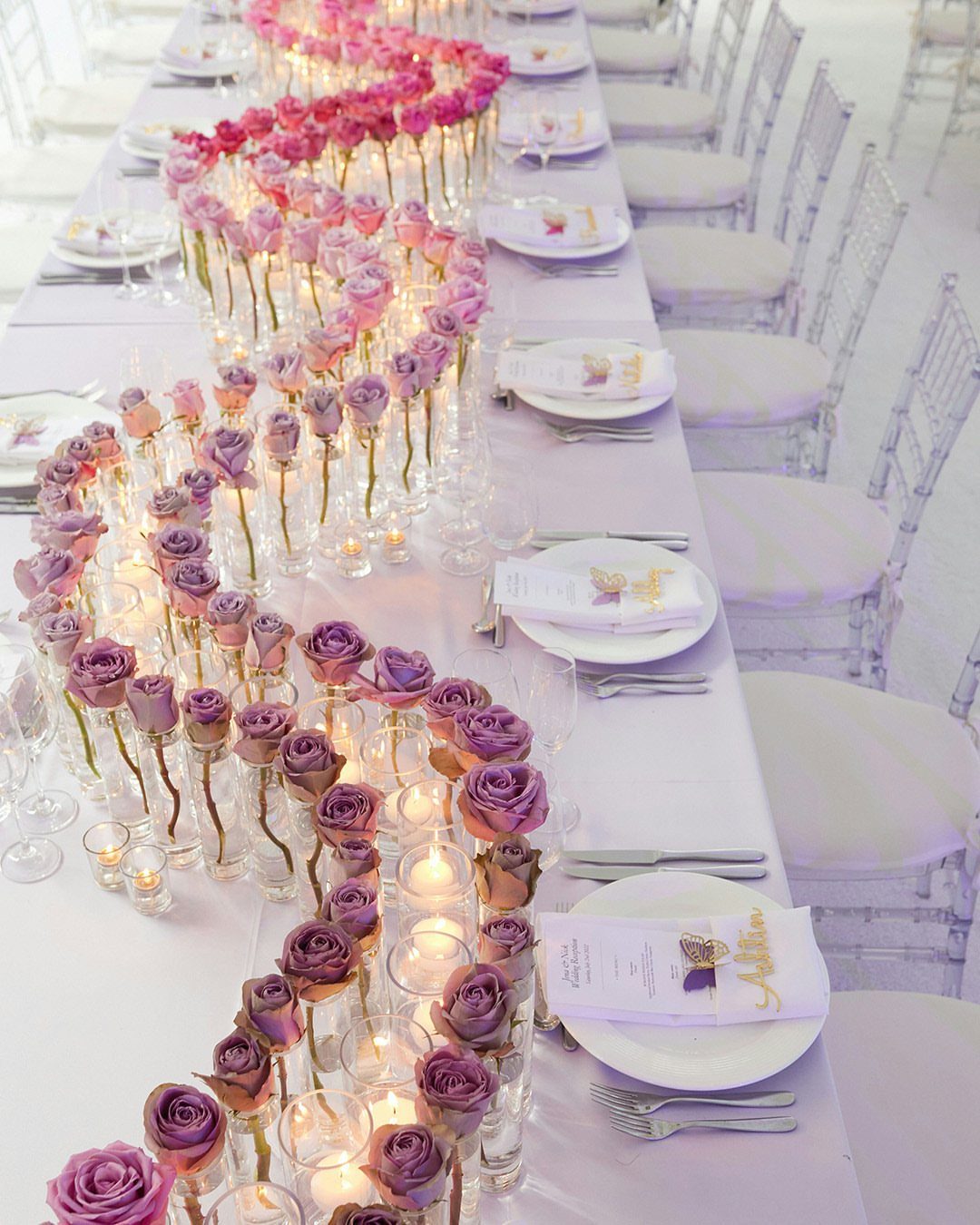 Pink and purple roses arranged in a winding gradient centerpiece with candles on a long white table.