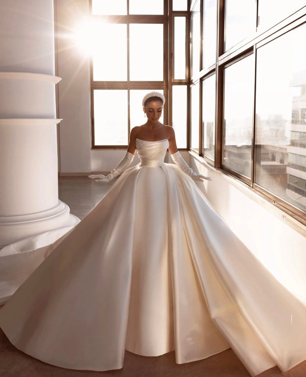 Woman in a voluminous white satin ball gown wedding dress standing by large windows.