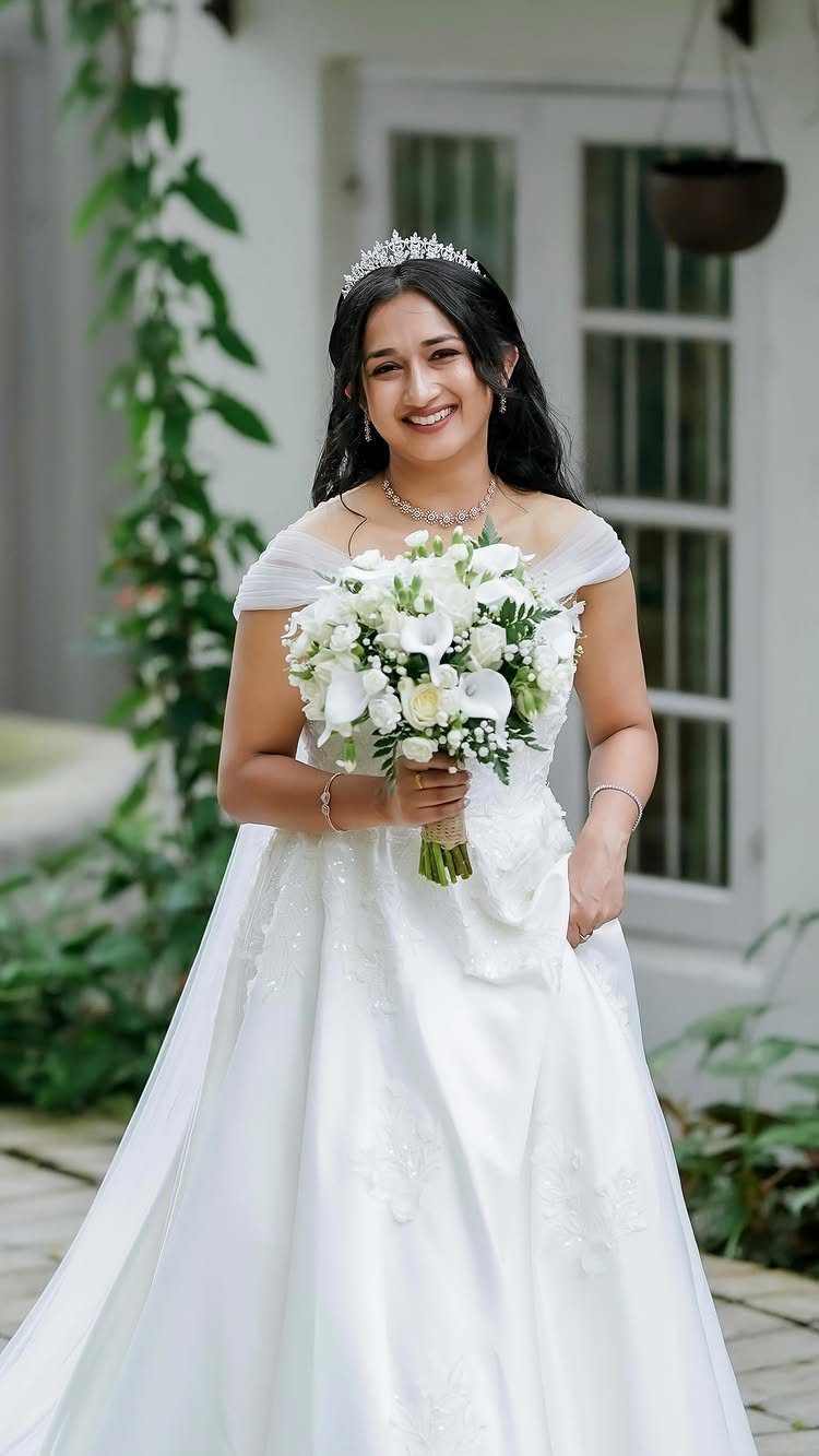 A smiling bride in an off-the-shoulder wedding dress holding a white flower bouquet.