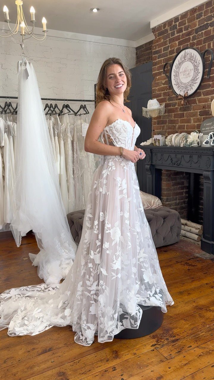 A smiling woman wearing a white floral lace sweetheart neckline wedding dress in a boutique.
