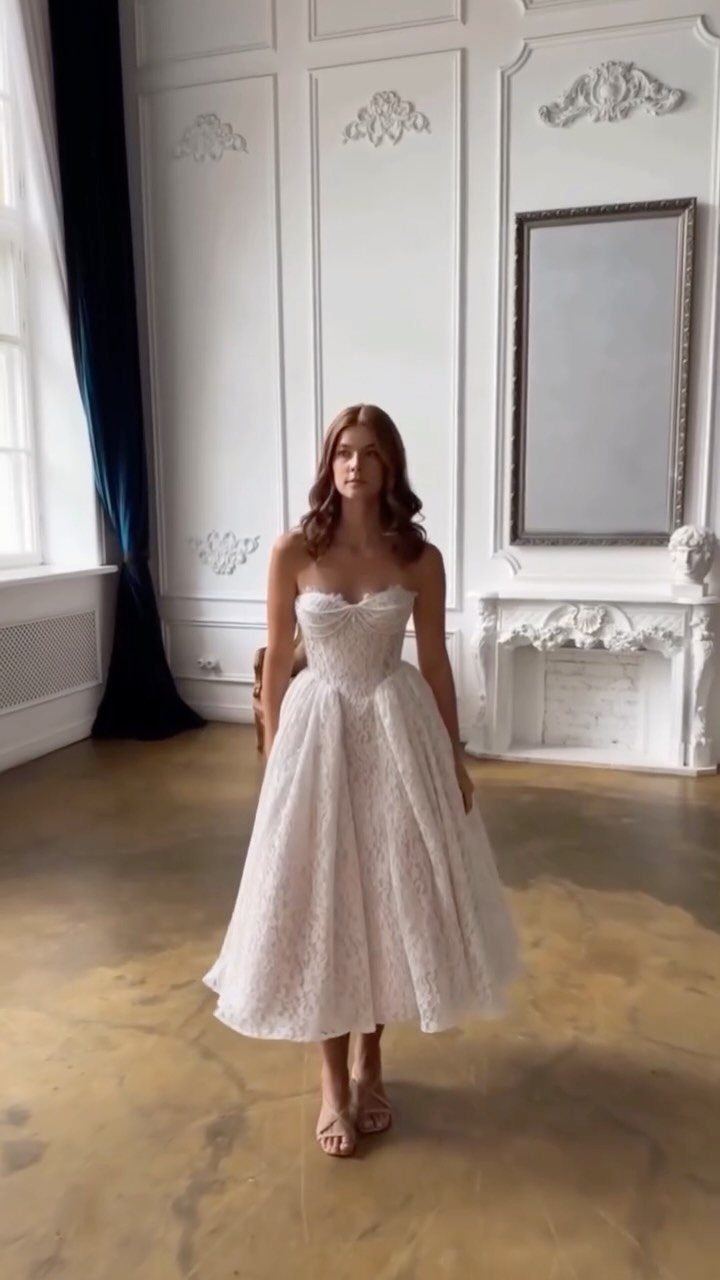 A woman wearing a strapless white lace tea-length wedding dress in a white room.