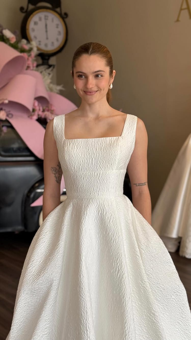 A smiling woman wearing a white textured wedding dress with a square neckline.
