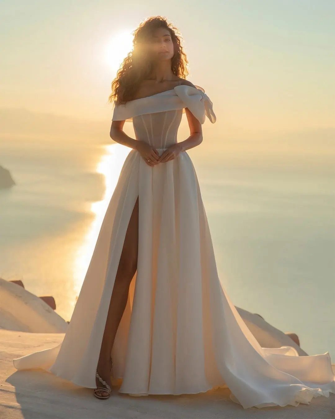 A woman in a white wedding dress with a high slit standing against a sunset ocean.