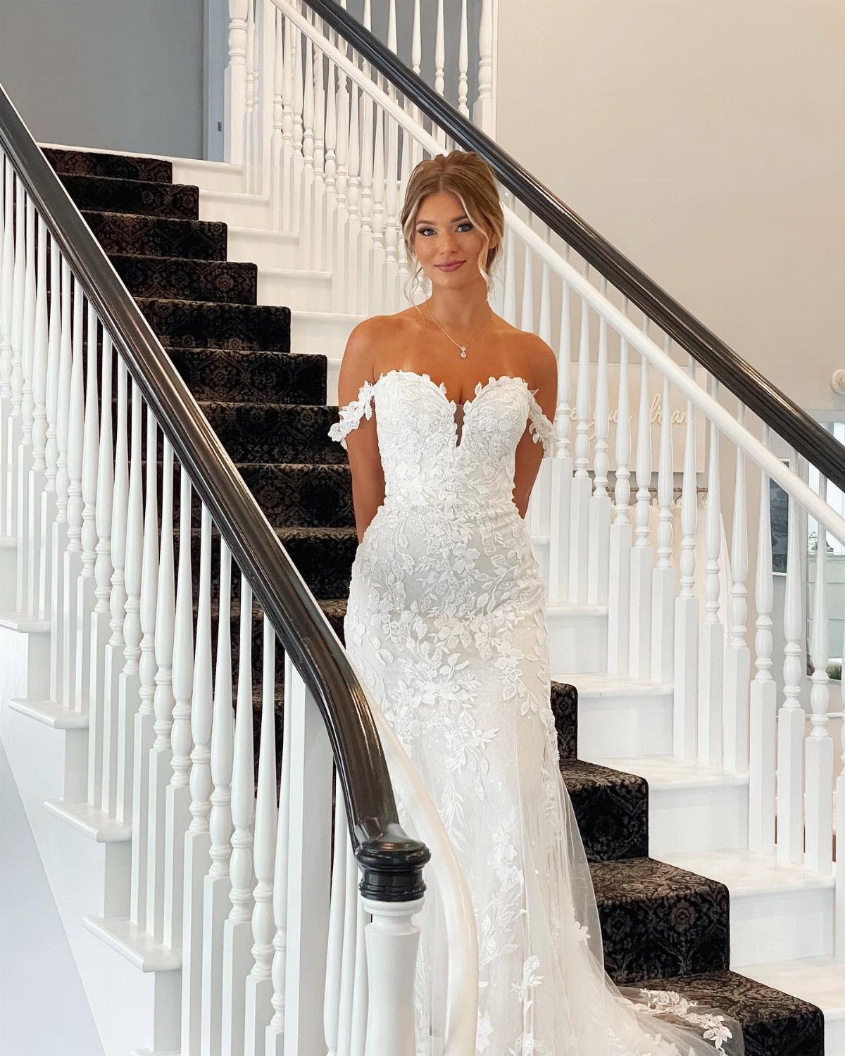 Woman wearing an elegant white lace off-the-shoulder wedding dress standing on a staircase.