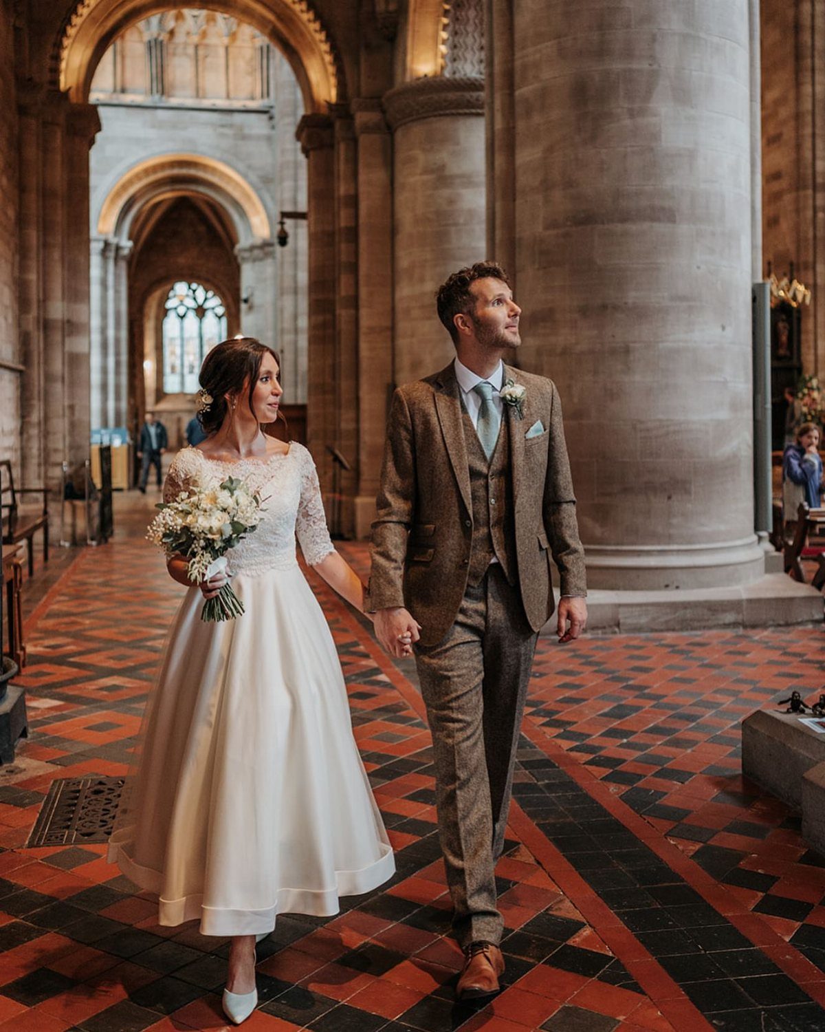 Bride in a white tea-length lace wedding dress walking with groom in a cathedral.