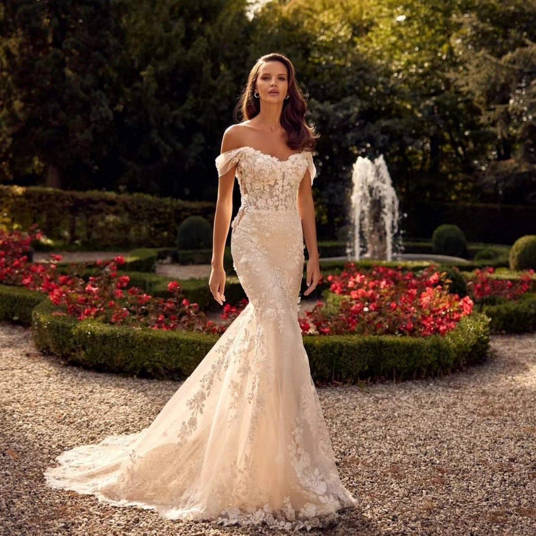 Woman wearing a lace trumpet wedding dress standing in a lush garden with a fountain.