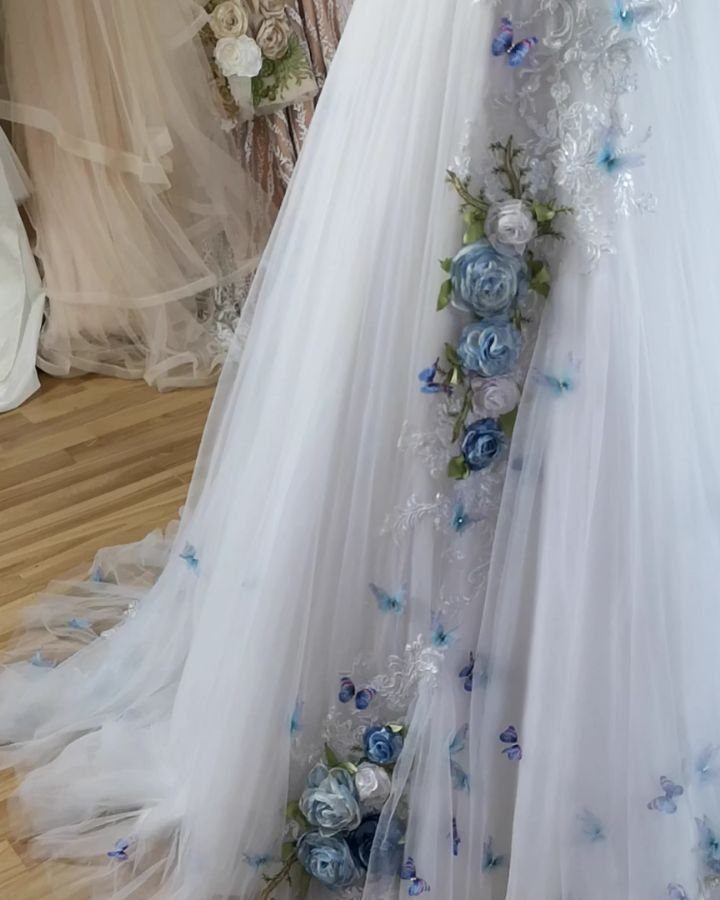 White wedding dress featuring blue fabric flowers and small blue butterfly decorations on tulle fabric.
