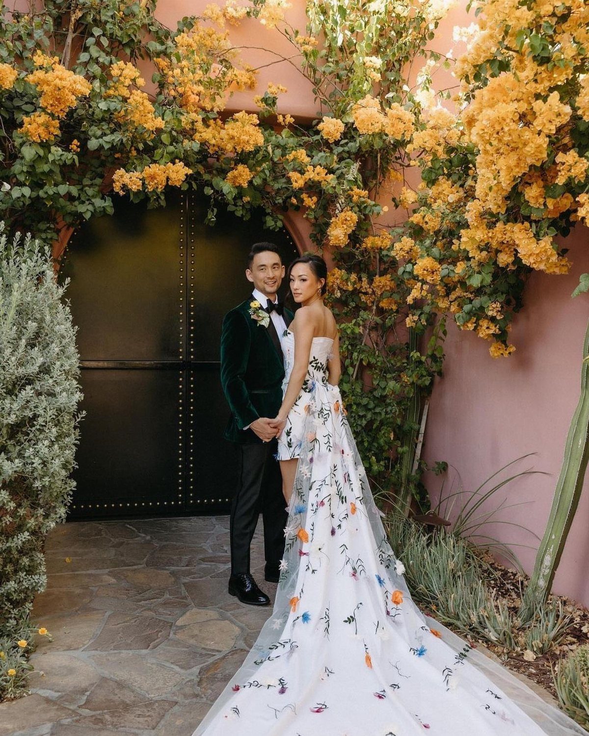 Bride in a white wedding dress with colorful floral appliqués posing with her groom outdoors.