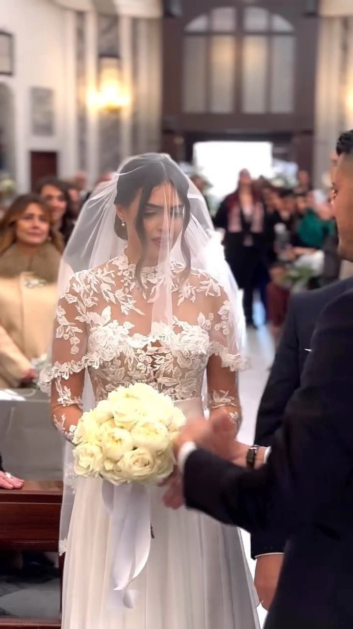 Bride in a lace wedding dress and veil walking down the aisle holding white roses.
