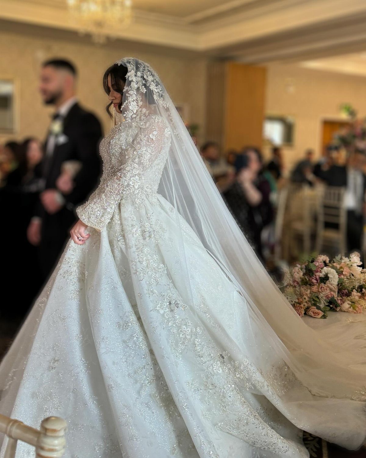 Bride in a lace wedding dress wearing a long, flowing sheer veil indoors.