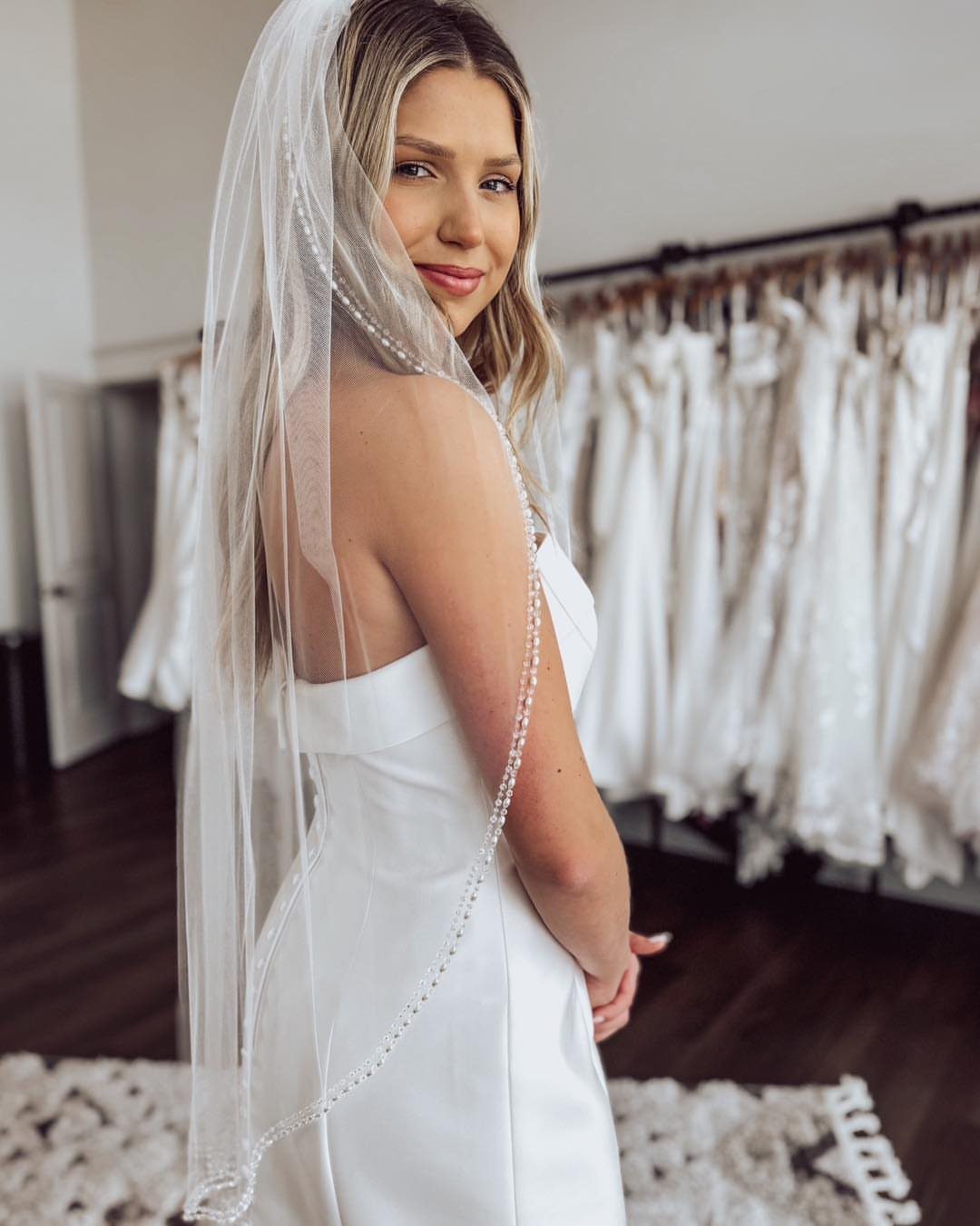 A woman wearing a white wedding dress and a beaded fingertip veil in a bridal shop.