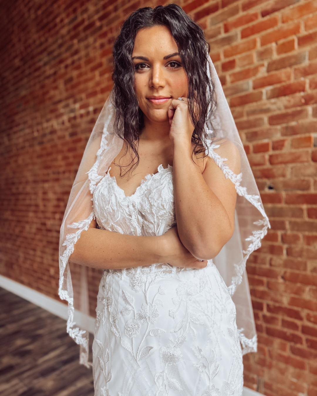 Woman in a white lace wedding dress and sheer veil posing against a brick wall.