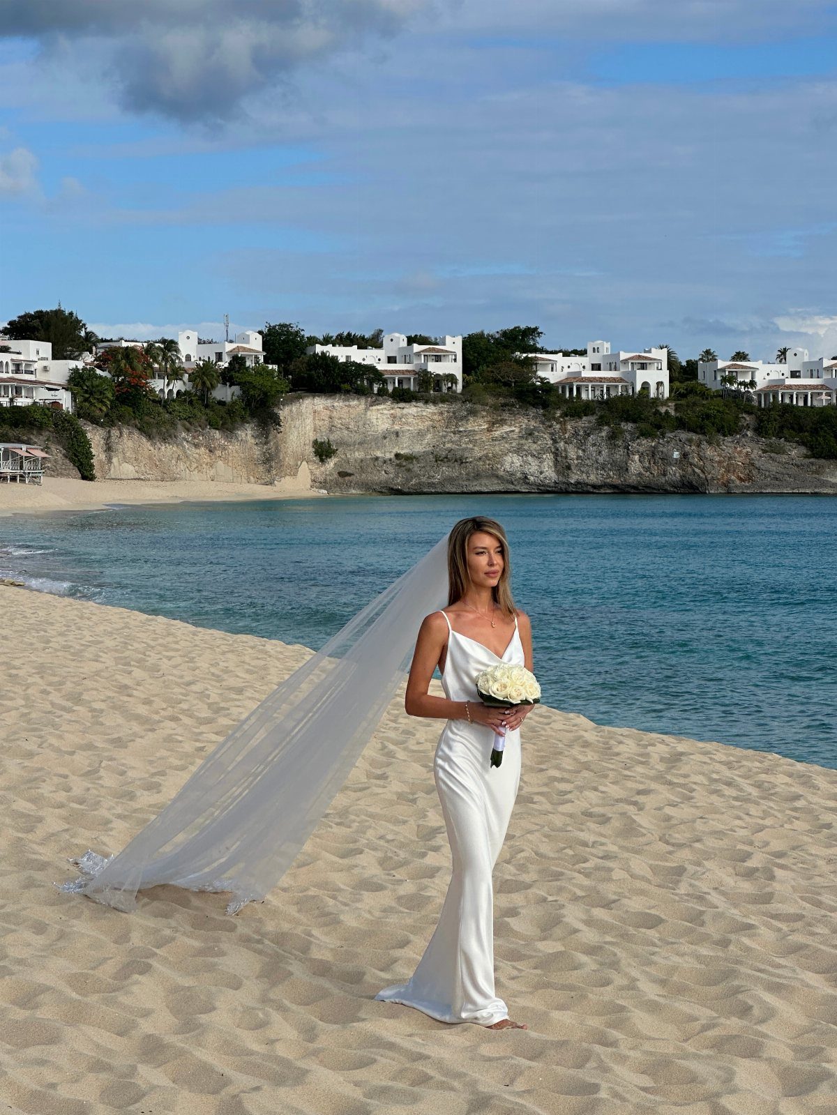 A woman in a white wedding dress and long veil standing on a sandy beach by the ocean.