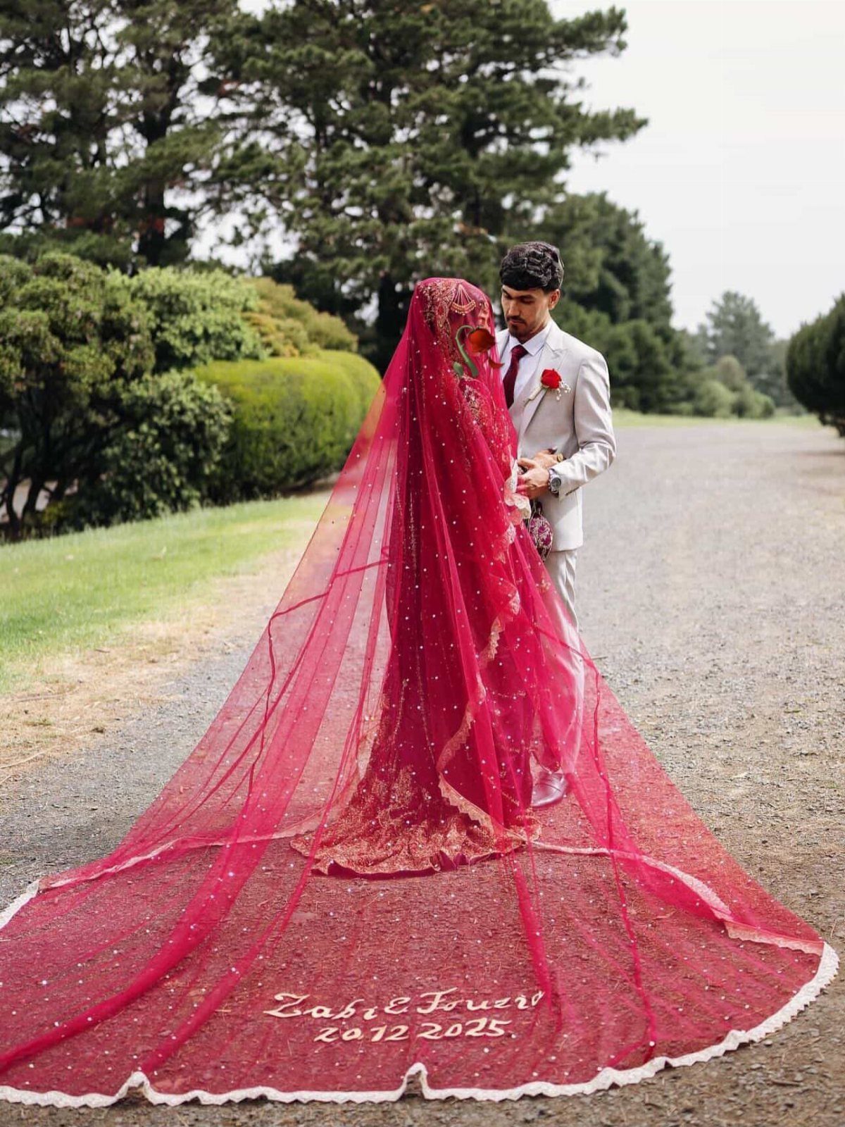A bride in a red dress and long red veil stands next to her groom outdoors.