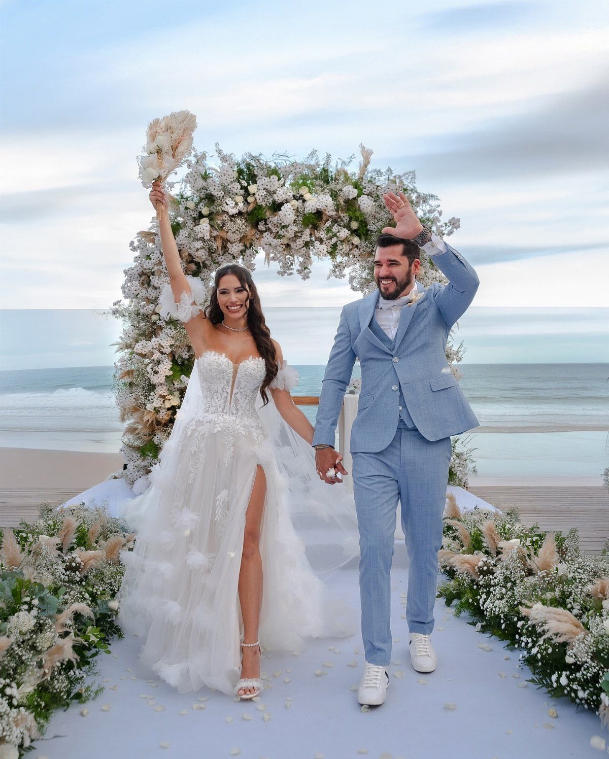 A bride in a white lace dress and a groom in a blue suit walking on a beach.