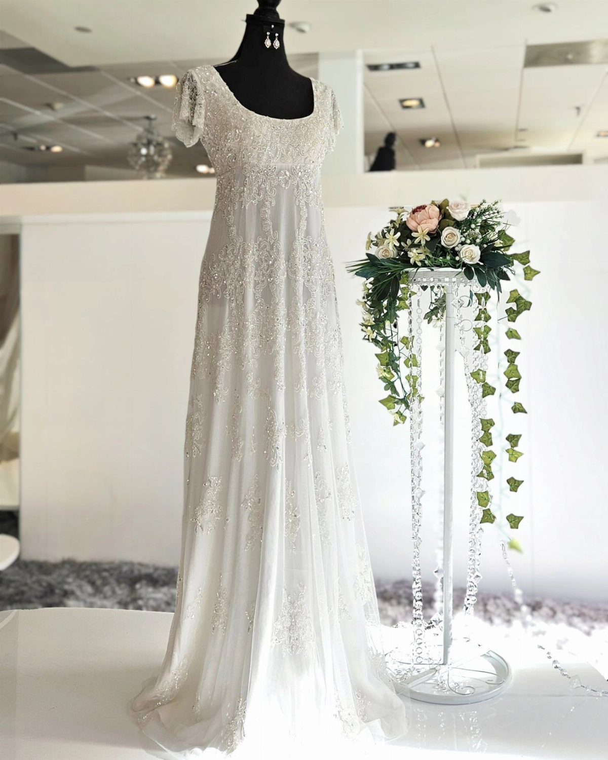White beaded wedding dress with short sleeves displayed on a mannequin next to flowers.