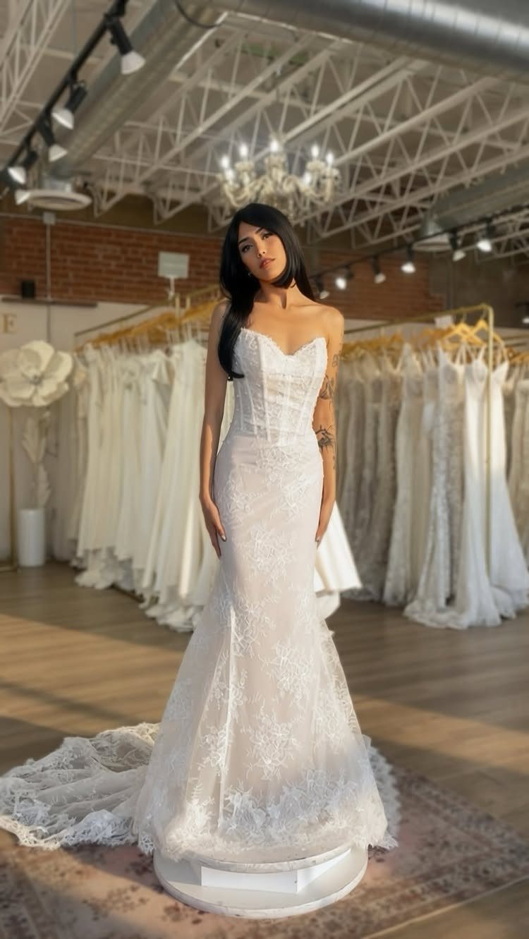 A woman posing in a white lace fit-and-flare wedding dress inside a bridal boutique.