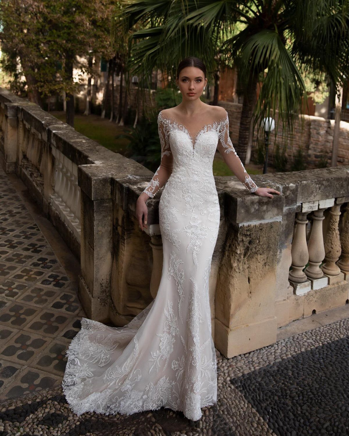 Woman wearing a long-sleeved lace mermaid style wedding dress standing by a stone railing.