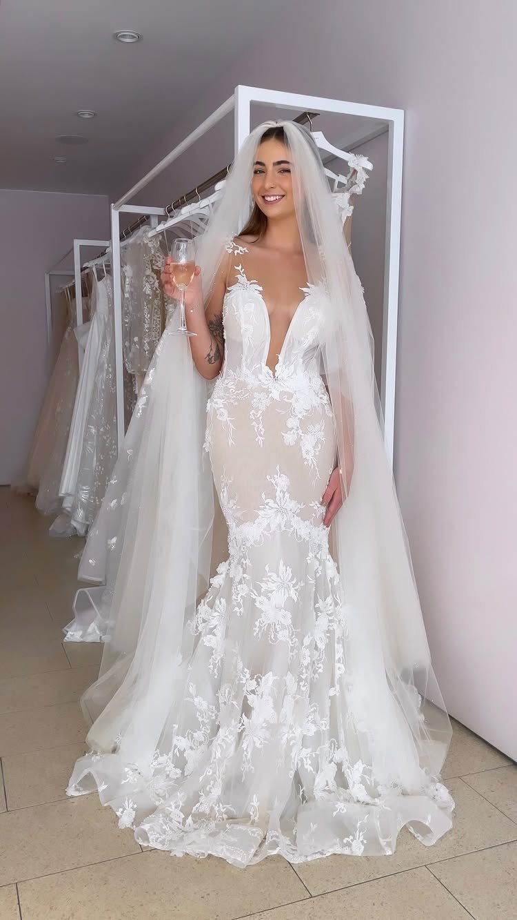 A smiling woman in a white lace wedding dress with a long veil holding a glass.