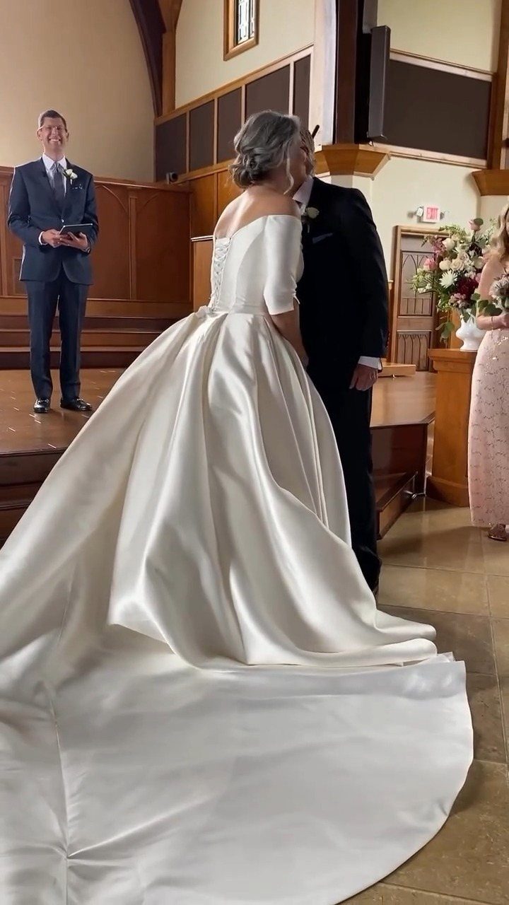 A bride wearing a white satin off-the-shoulder ballgown with a long train stands indoors.