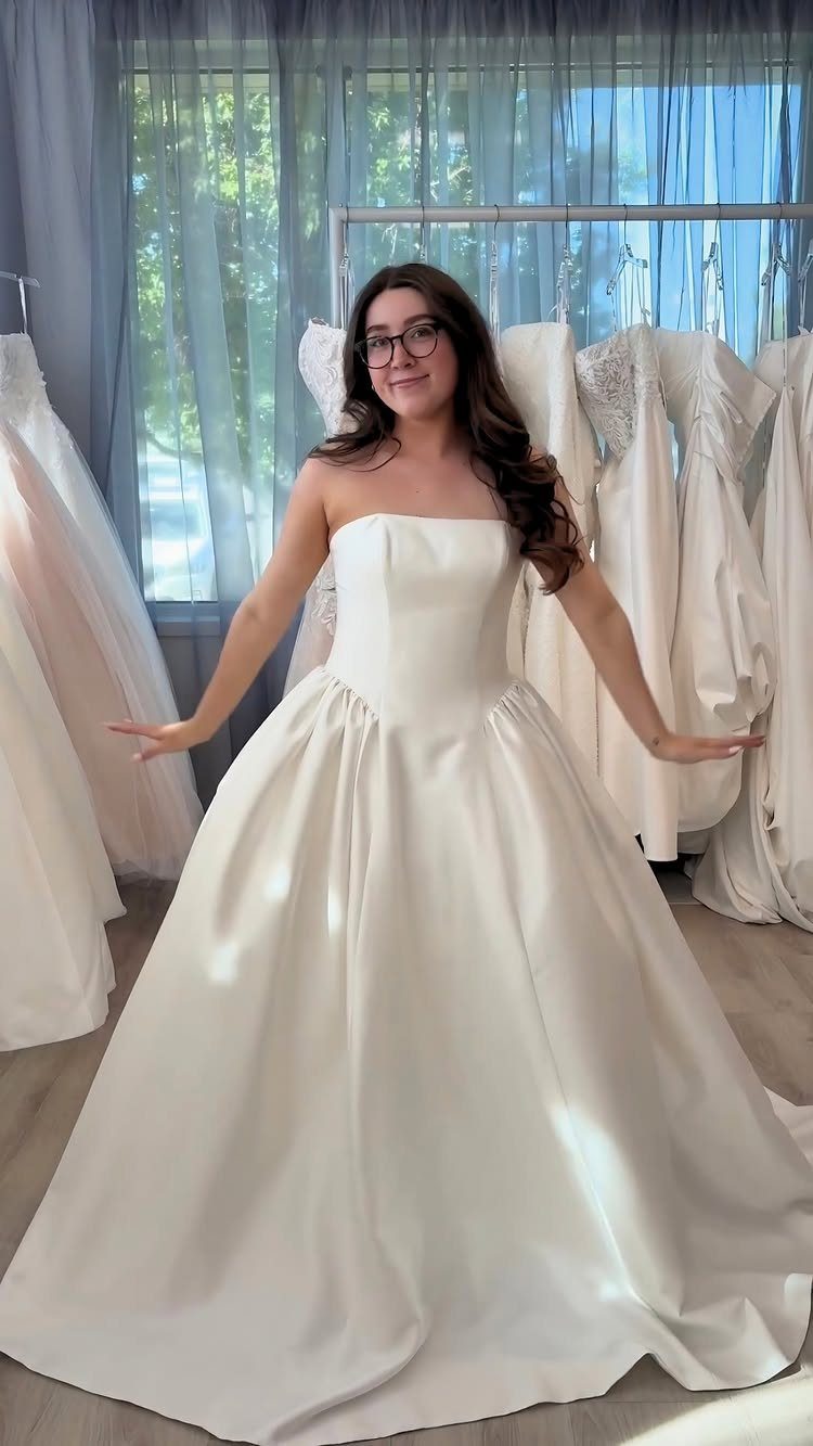 A smiling woman posing in a strapless white satin wedding gown inside a bridal boutique.