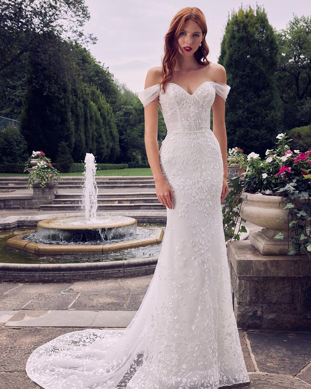 A woman in an off-the-shoulder white lace wedding dress standing in a garden by a fountain.