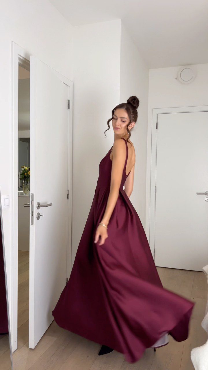 Woman twirling in a long, flowing burgundy satin sleeveless gown in a bright room.