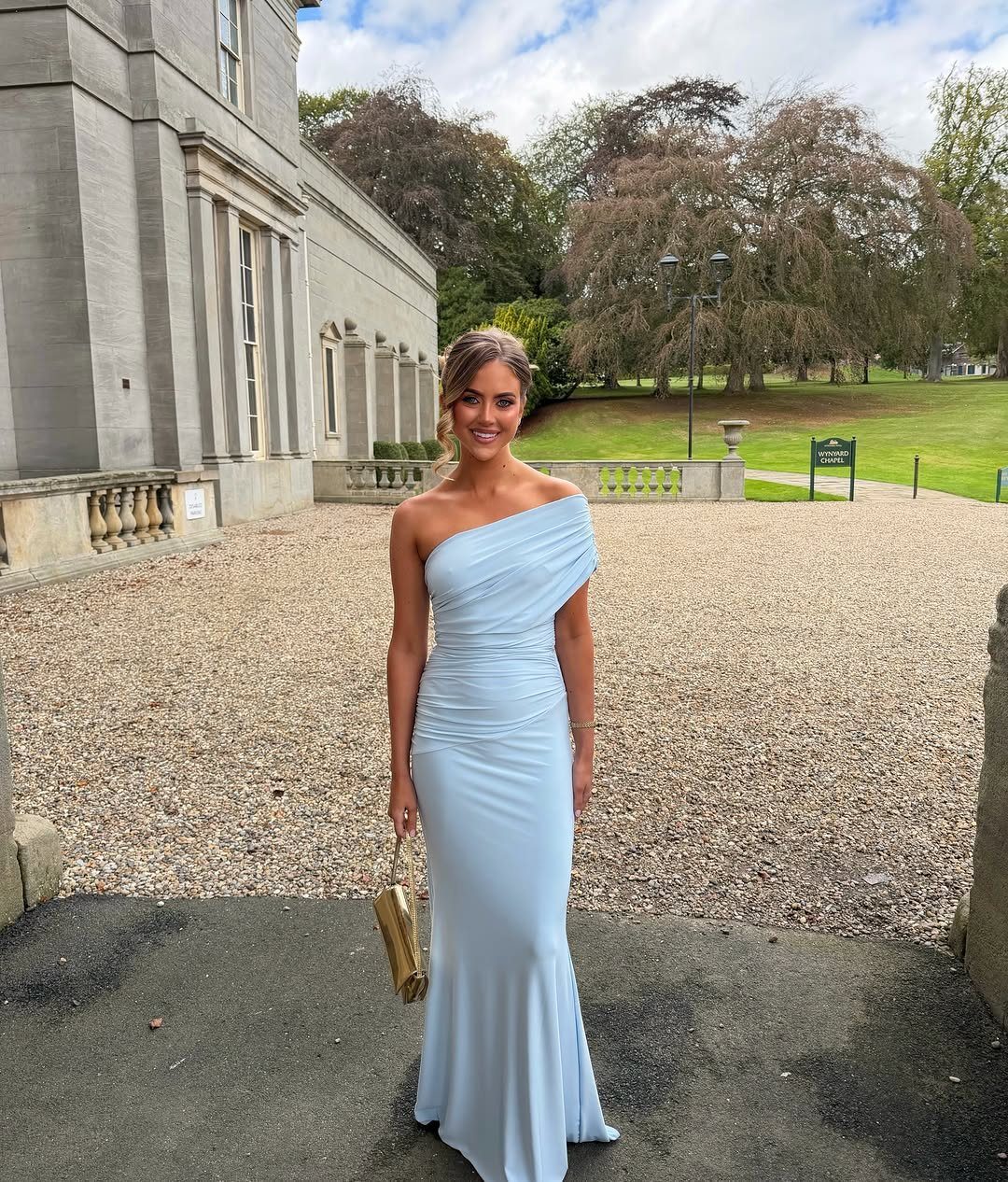 A woman posing in a light ice blue one-shoulder mermaid gown holding a gold clutch.
