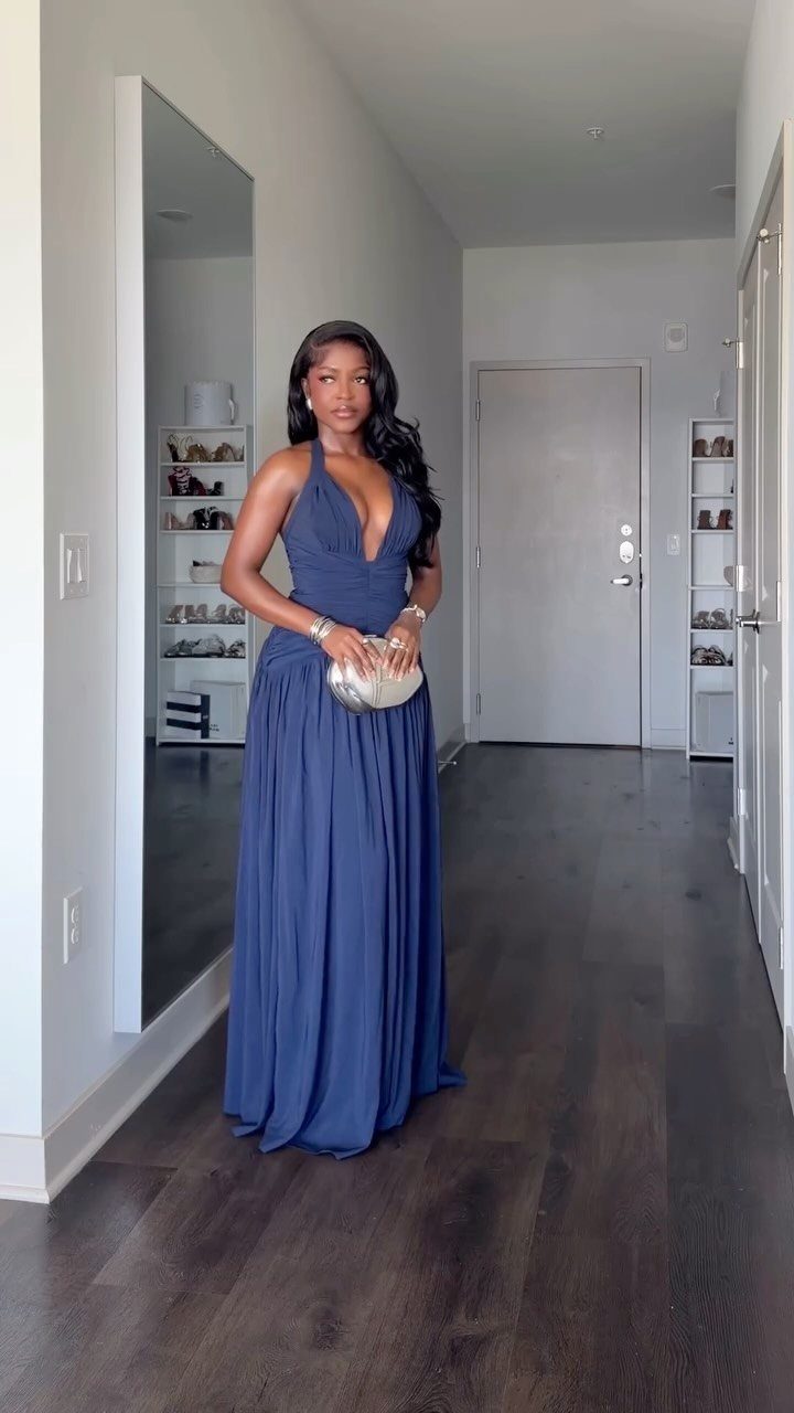 A woman posing in a long, deep blue halter maxi dress and holding a silver clutch.